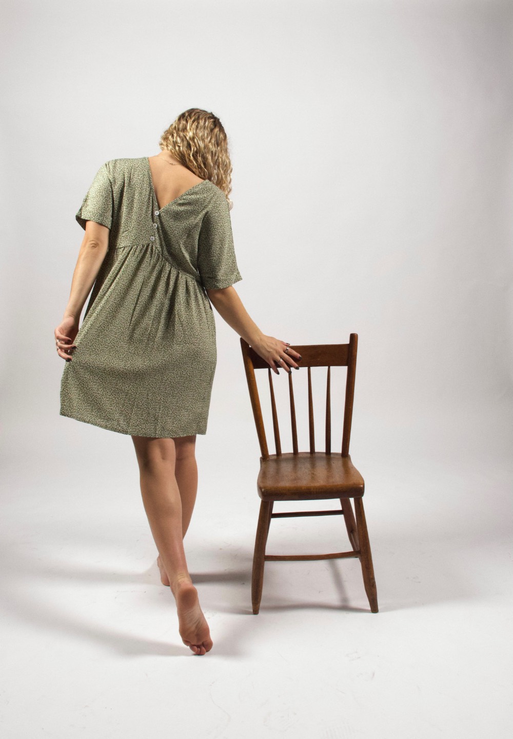 The back of a woman in a green dress with one hand on a wooden chair.